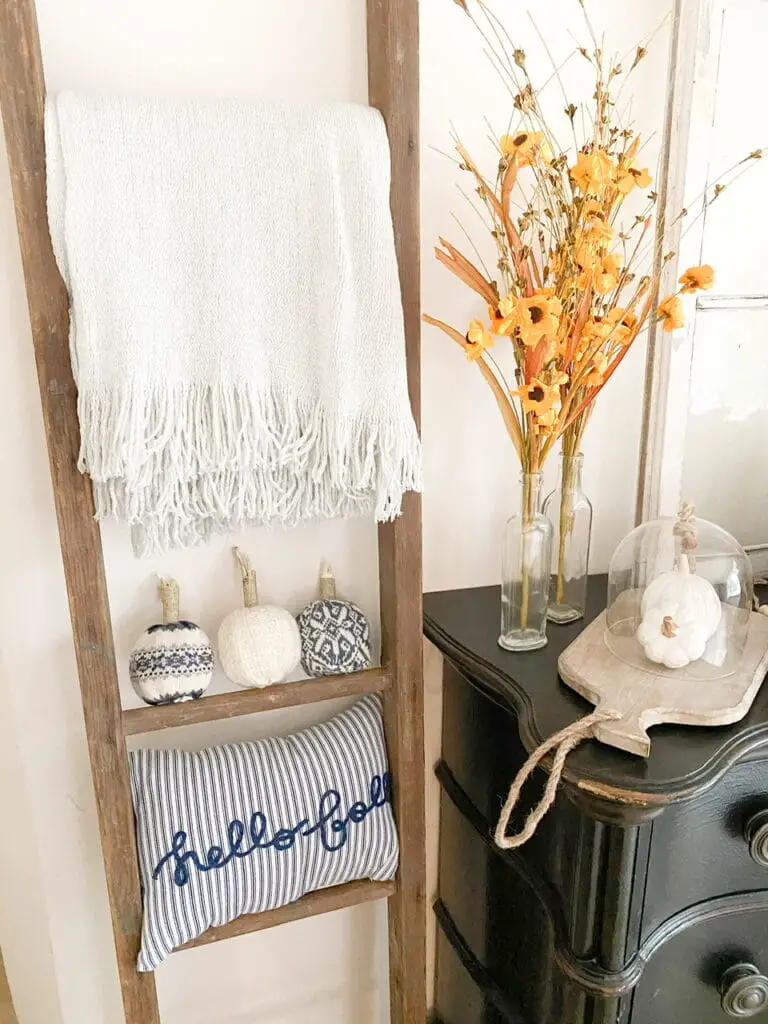 Photo shows the 3 completed DIY sweater pumpkins sitting on an antique ladder underneath a grey throw blanket and above a blue and white striped pillow that says "hello fall" It also shows a black antique looking dresser with some yellow paper flowers in some clear bottle vases, a wood tray with 2 ceramic white pumpkins underneath a glass cloche.