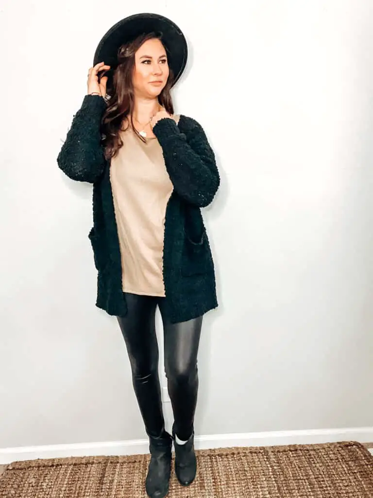 This thrifted winter outfit is made up of a camel color sweater underneath a textured knit black cardigan, paired with some black faux leggings, a black hat, a gold medallion necklace, and some black faux leather ankle boots.