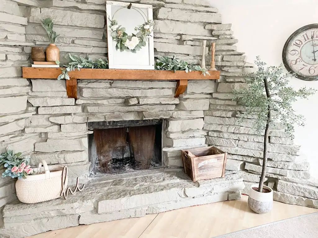 A spring home tour featuring vintage and vintage inspired pieces as well as some thrifted home decor items. This image shows a large gray stone fireplace with a wood mantel decorated with a garland, a window mirror, a spring hoop wreath, some wooden candle holders, some wooden vases on top of old books, a large white washed woven basket with a mixed floral and greenery bouquet sticking out the top, a wood crate box, and an artificial tree.