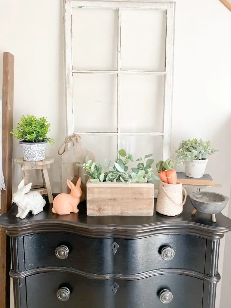 This image shows a black entryway table with an antique window, various spring decor, some small potted plants, and some wood decor items.
