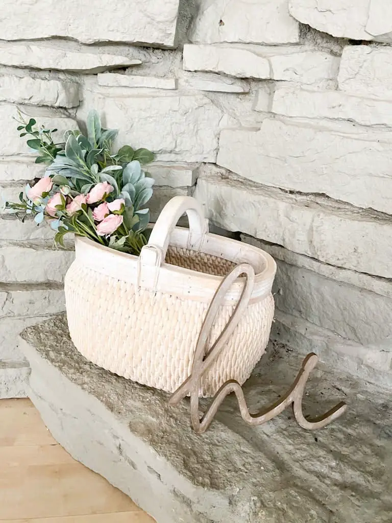 A large whitewashed basket with a wooden handle over top that has a mixed bouquet of pink flowers and greenery inside, next to that is a wooden "hi."