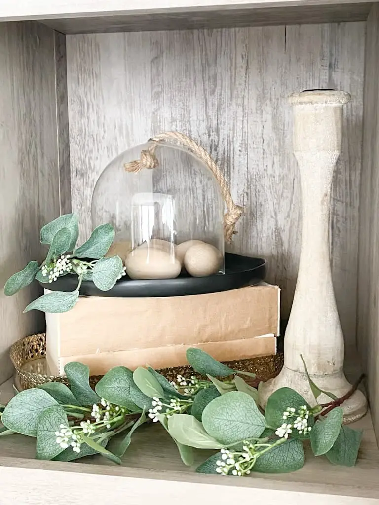 A stack of thick books with no covers on them on top of a mirrored tray with a glass cloche housing some wooden eggs on top of a black ceramic plate, next to that is a wood candlestick holder and there is seeded eucalyptus spread throughout.