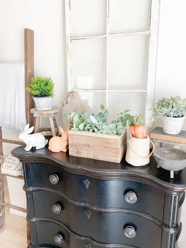 This image shows a black entryway table with an antique window, various spring decor, some small potted plants, and some wood decor items.