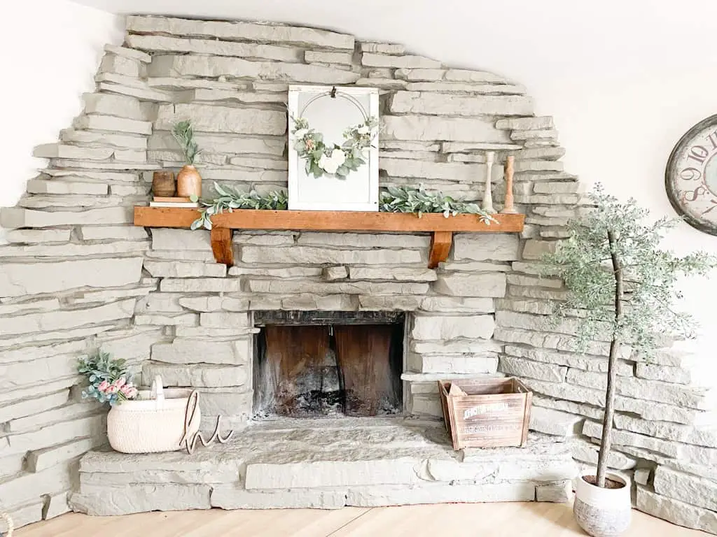 This image shows a large gray stone fireplace with a wood mantel decorated with a garland, a window mirror, a spring hoop wreath, some wooden candle holders, some wooden vases on top of old books, a large white washed woven basket with a mixed floral and greenery bouquet sticking out the top, a wood crate box, and an artificial tree.