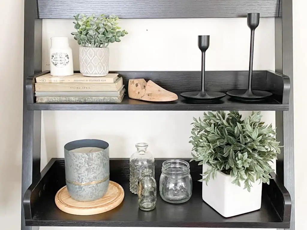 A two-tiered black wall shelf that has a stack of old books with an onld English advertising pot and a small potted plant on top, an old wooden shoe form, 2 black metal candlestick holders, a metal candle on top of a circular wood tray, 3 assorted small glass jars, and a plant in a white ceramic pot.