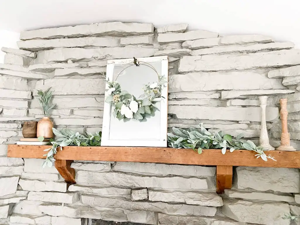 This image shows a gray stone fireplace with a wood mantel decorated with a garland, a window mirror, a spring hoop wreath, some wooden candle holders, and some wooden vases on top of old books.