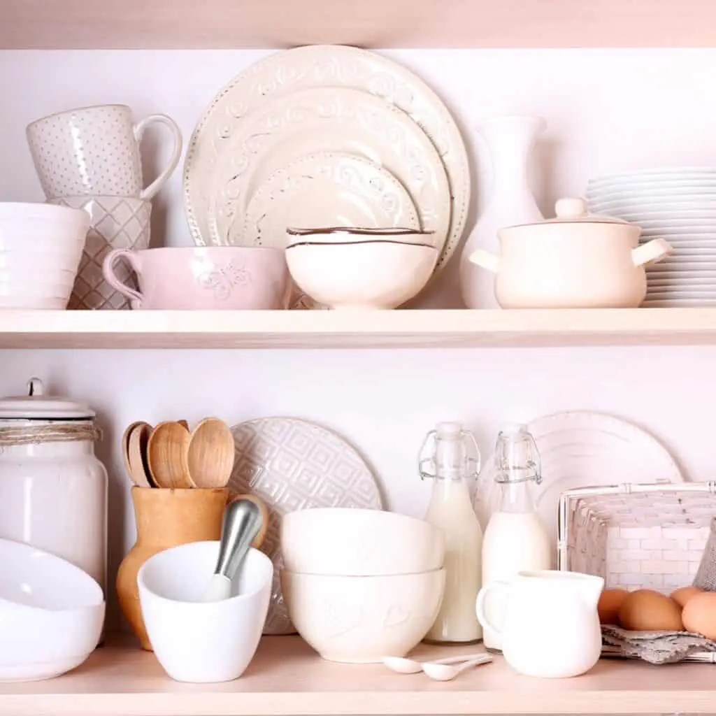 Light wood shelves with a variety of white tableware and a mixture of wood and white kitchen items. There are also some glass kitchen items mixed in.