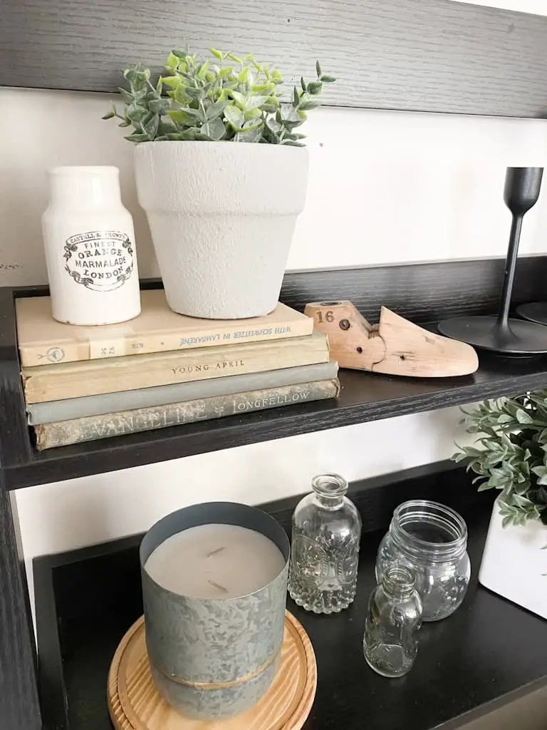 A light gray textured pot with a small plant inside next to an antique advertising jar, both are on top of a stack of old books. Next to that is an old wooden shoe form and below it are a candle in a metal holder on top of a round wood circle and some antique glass jars.