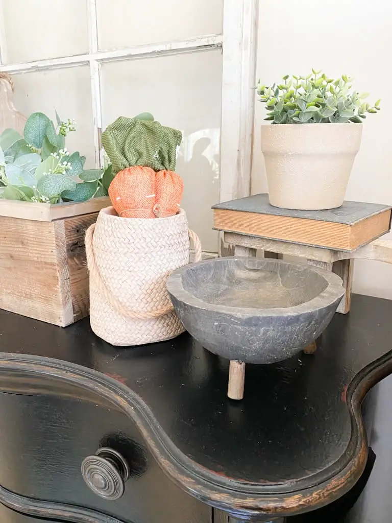 A tan textured pot with a small green plant inside on top of a book and a small stool. In front of that is a weathered black wooden bowl with legs next to a woven pottery vase with 3 fabric carrots sticking out the top of it.