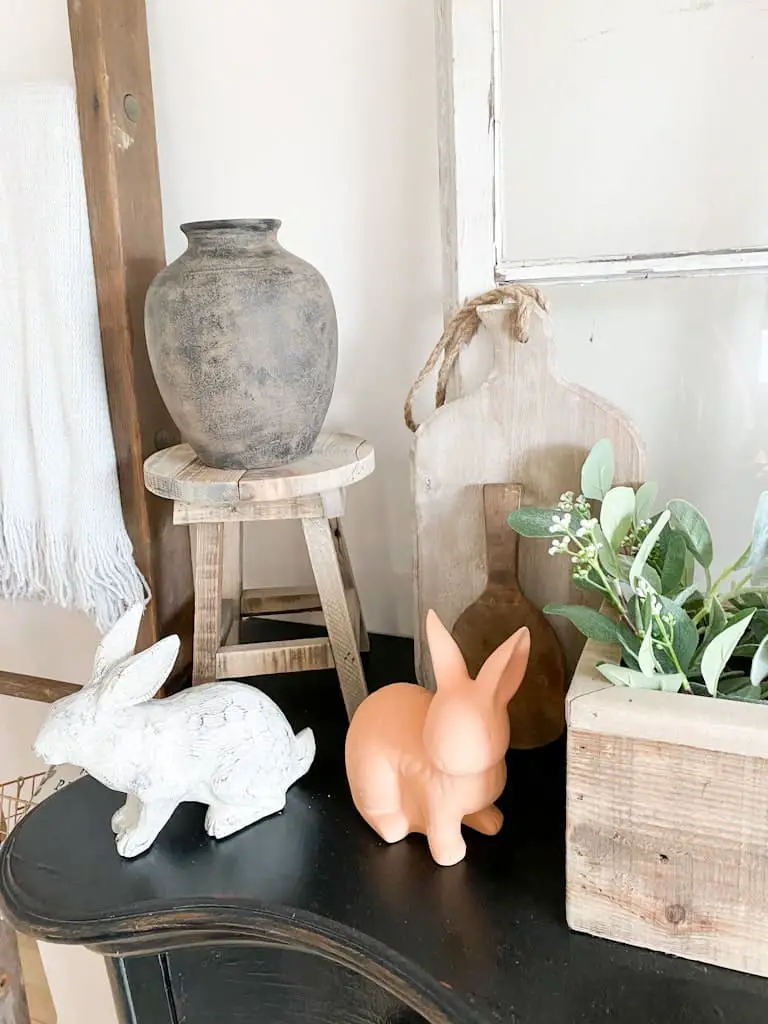 The upcycled aged pottery vase after it was finished, sitting on an old stool with a white wood bunny and a terra cotta bunny sitting in front of it.