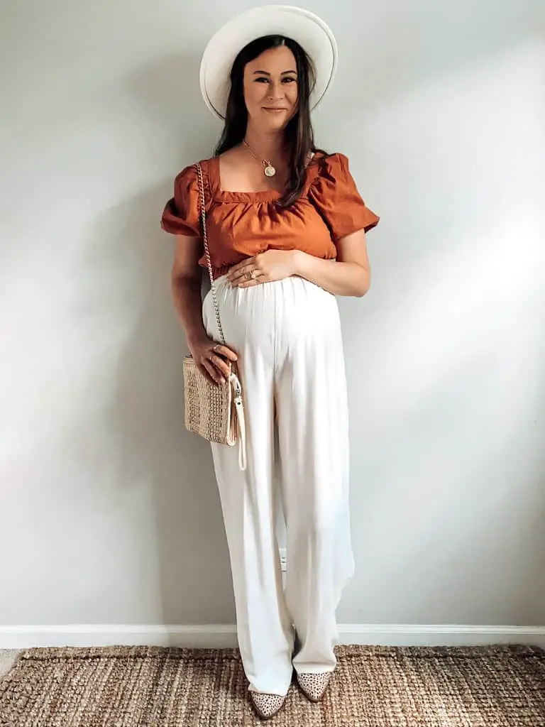 A pregnant woman wearing a rust colored cropped shirt with some high rise cream casual trousers, a wide brim hat that is cream, a straw bag with a gold chain, and some leopard print boots.