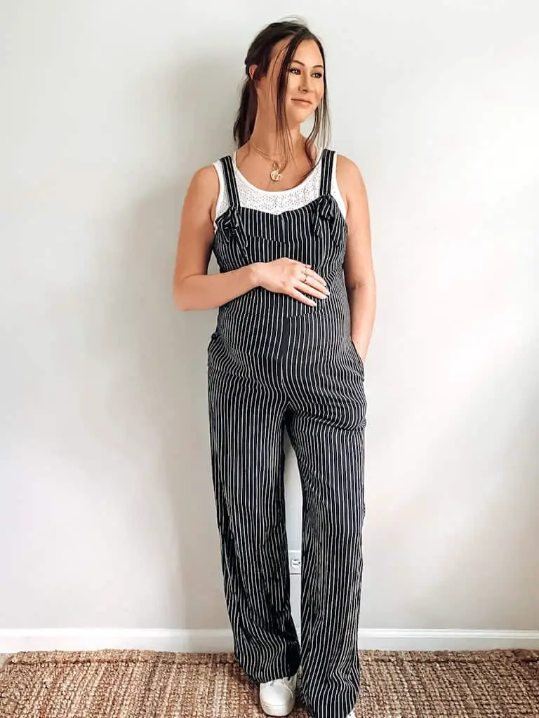 A pregnant woman wearing black and white striped overalls with a white eyelet tank underneath, some gold necklaces, and white sneakers. These are both maternity second hand clothes that are actually regular clothes you can wear while pregnant.