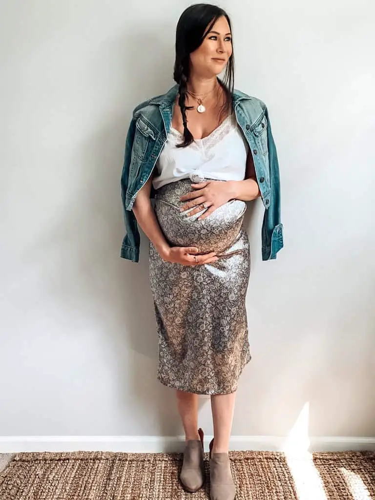 A pregnant woman wearing a white lace trim camisole, a lavender satin midi skirt, a denim jacket draped over the shoulders, some gold necklaces and taupe ankle boots.