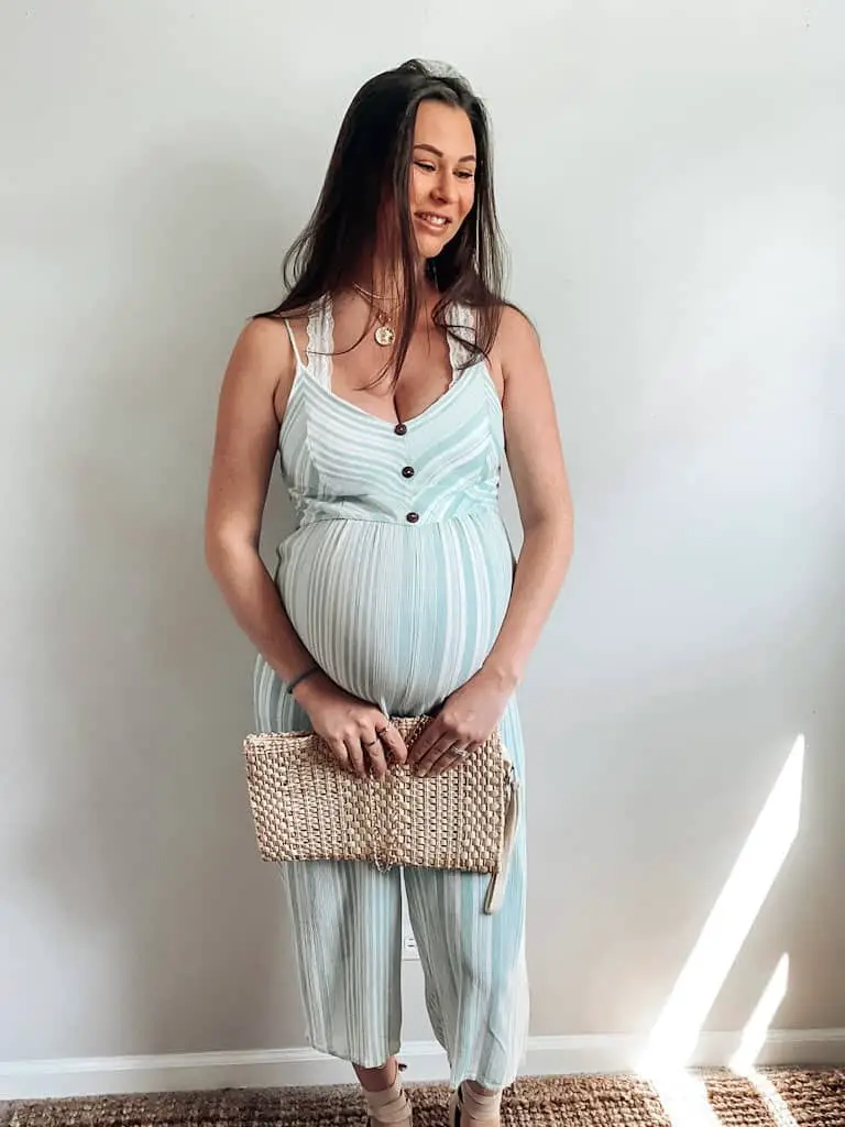 A pregnant woman wearing a green and white striped romper with faux wood buttons, holding a straw clutch, wearing some tan espadrilles and some gold necklaces.
