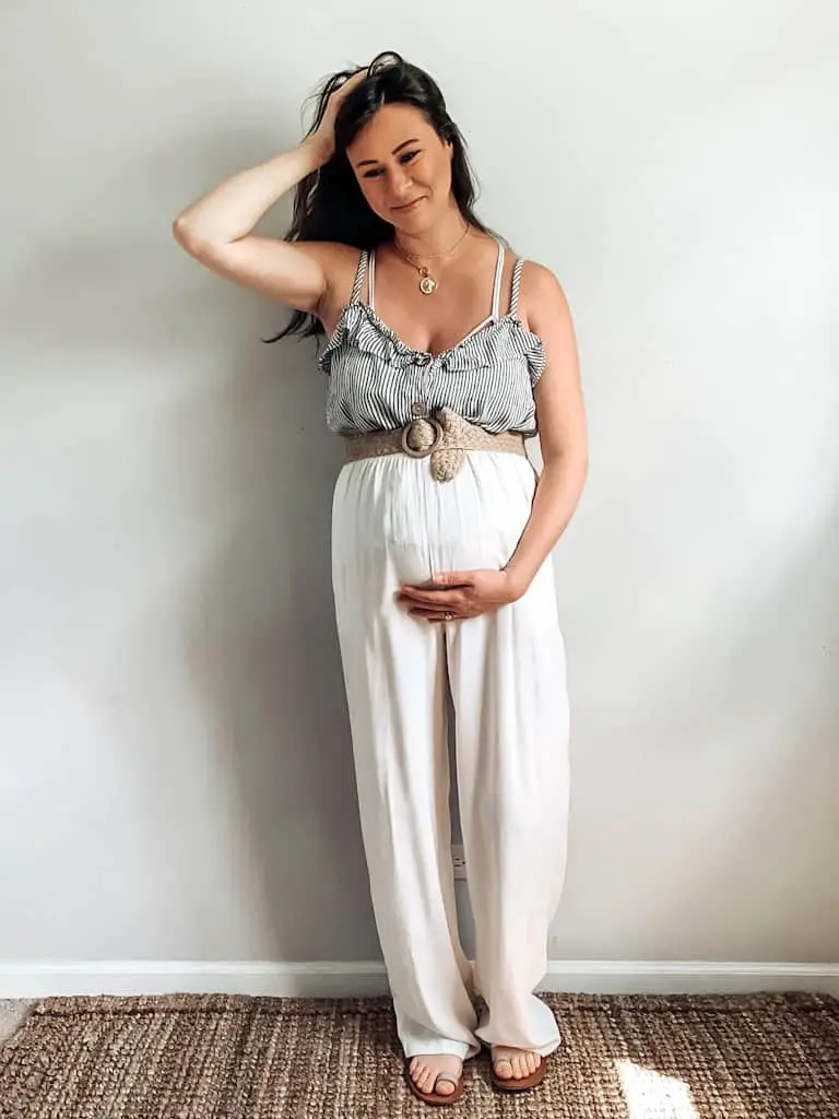A pregnant woman wearing a blue and white striped top with a ruffle neckline and wooden buttons, cream wide leg high rise casual trousers, a tan woven belt, some gold necklaces, and sandals. These are both maternity second hand clothes that are actually regular clothes you can wear while pregnant.