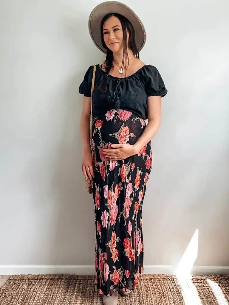 A pregnant woman wearing a black eyelet cropped shirt, a black maxi skirt with pink and purple flowers on it, a gray wide brim hat, a straw bag, gold necklaces, and taupe ankle boots. These are both maternity second hand clothes that are actually regular clothes you can wear while pregnant.