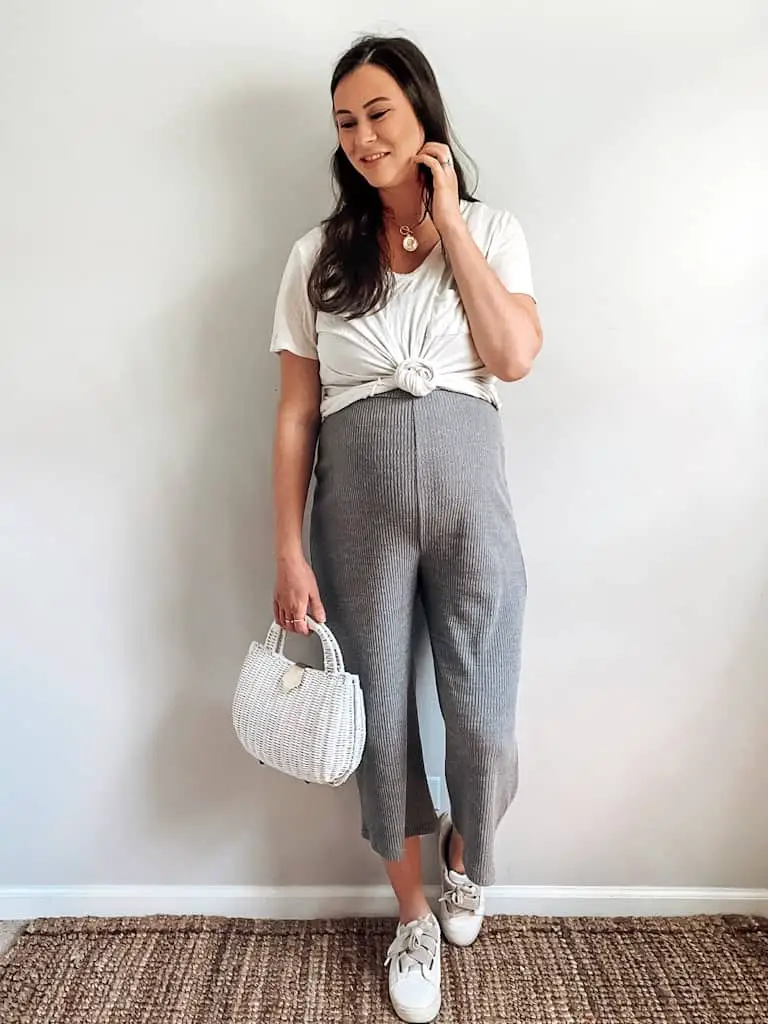 A pregnant woman wearing a gray ribbed jumpsuit with a white t-shirt over it that is knotted in front, a white wicker bag, some gold necklaces, and white sneakers.