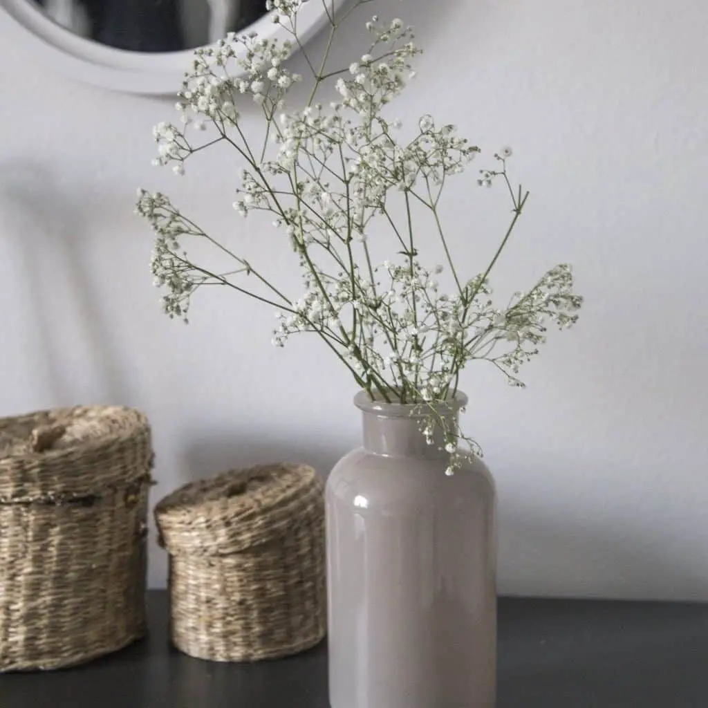 Some baby's breath in a warm tone gray vase next to 2 wicker containers with lids sits on a black table in front of a white wall and there is the edge of a white framed round mirror showing at the top of the photo.