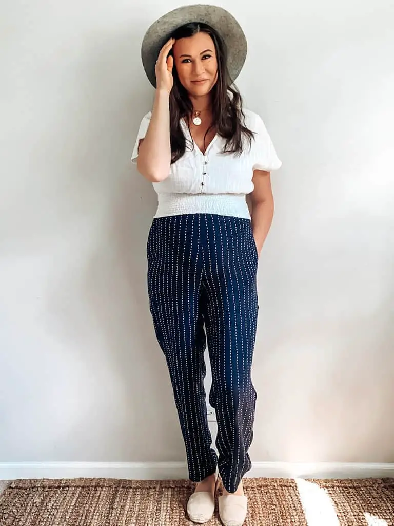 A pregnant woman wearing a white cropped shirt with buttons down the front and a gathered waist, blue and white striped high rise casual trousers, an oatmeal colored wide brim hat, gold necklaces, and cream espadrilles. These are both maternity second hand clothes that are actually regular clothes you can wear while pregnant.
