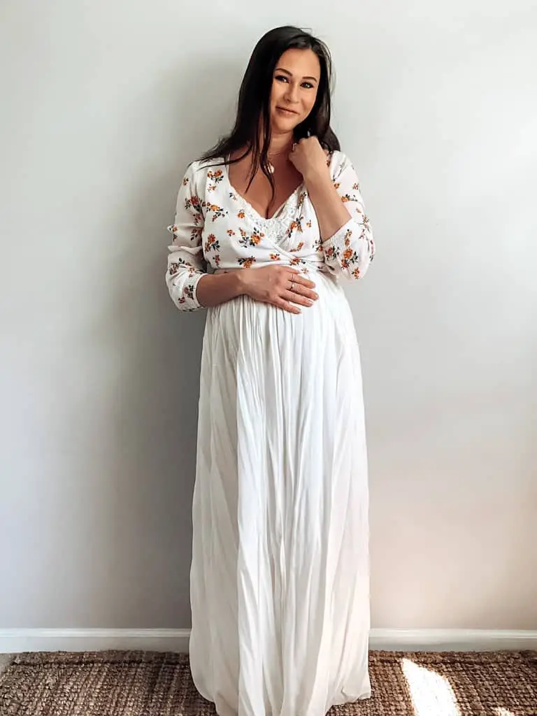 A pregnant woman wearing a white top with orange flowers over a white flowy maxi dress, and some gold necklaces. Maxi dresses are a perfect example of how to dress during pregnancy without maternity clothes.