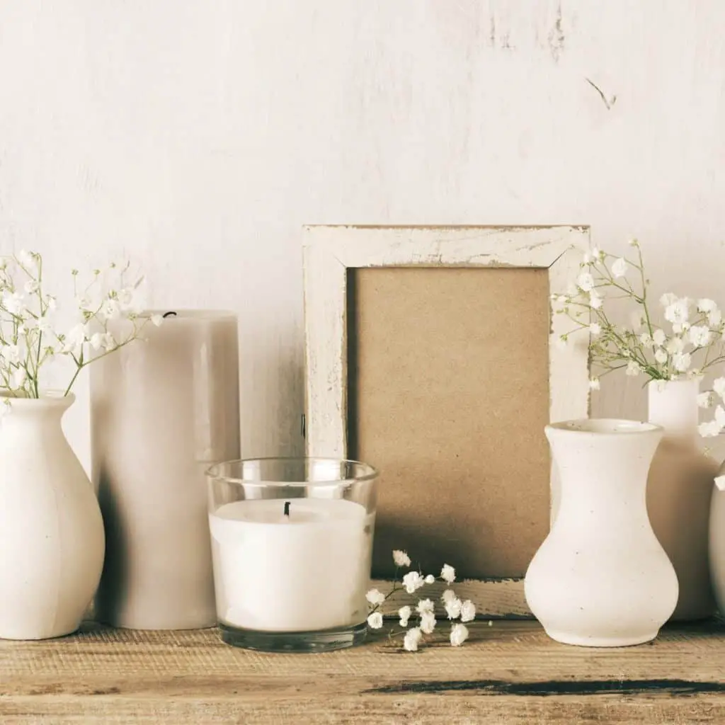 White vases with babay's breath in them, a gray candle, a white candle in a glass jar, an empty white distressed wood frame, all sitting on a weathered wood bench.