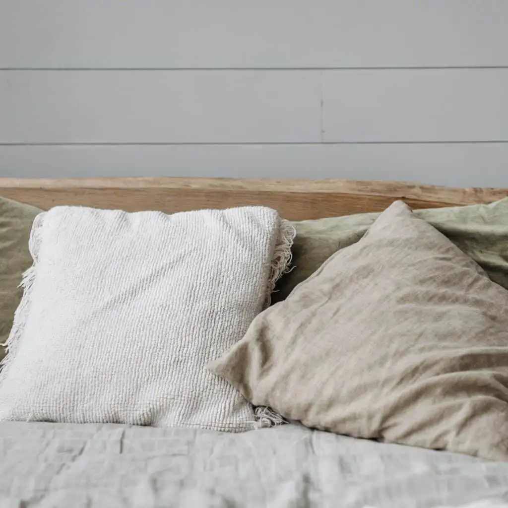 Adding neutral colored textiles will make your home oozy like this set of pillows which are white, tan, and light sage green and are a linen material. They are on top of a white linen comforter on a bed with a weathered wood headboard. The bed is in front of a white shiplap wall.