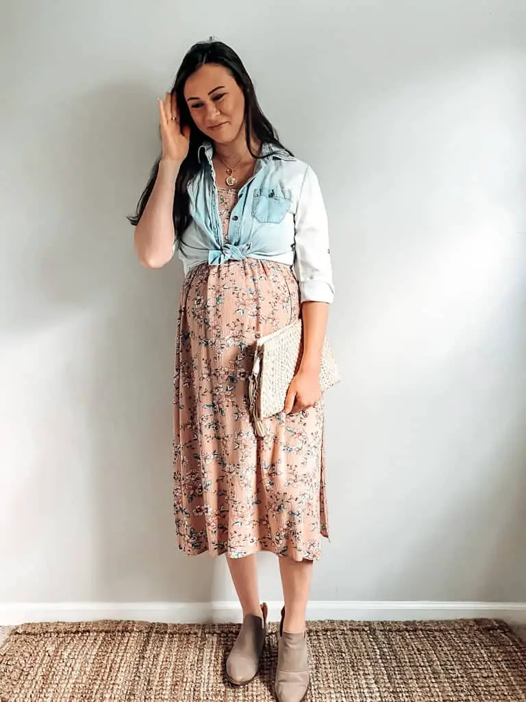 A pregnant woman wearing a pink floral midi length dress with a chambray button down shirt worn open and tied at the bottom over top of the dress, a straw handbag, some gold necklaces, and taupe ankle boots.