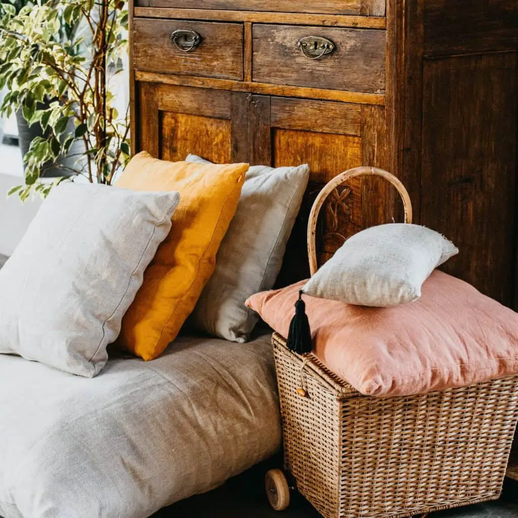 Some cream pillows mixed with a marigold and soft pink pillow which is in a wicker basket with a handle next to a couch which is in front of an antique cabinet and there is a potted houseplant next to it.