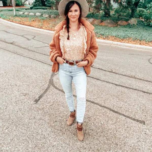 A woman standing outside wearing a gray wide brim hat, a yellow floral top under a brown teddy bear style jacket, some light wash high rise jeans with a belt, and brown ankle boots.