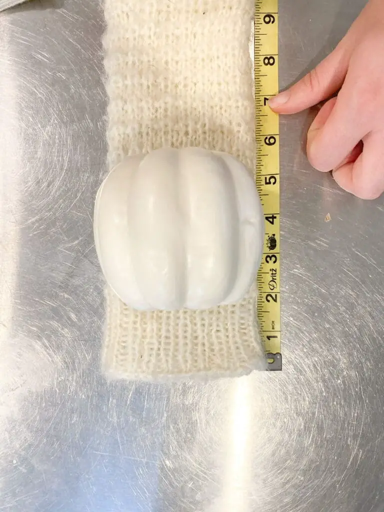 Photo shows the small white foam pumpkin laying on the cream glove next to a measuring tape with my finger pointing at the 7" mark. This is how much material I used for my DIY sweater pumpkin.