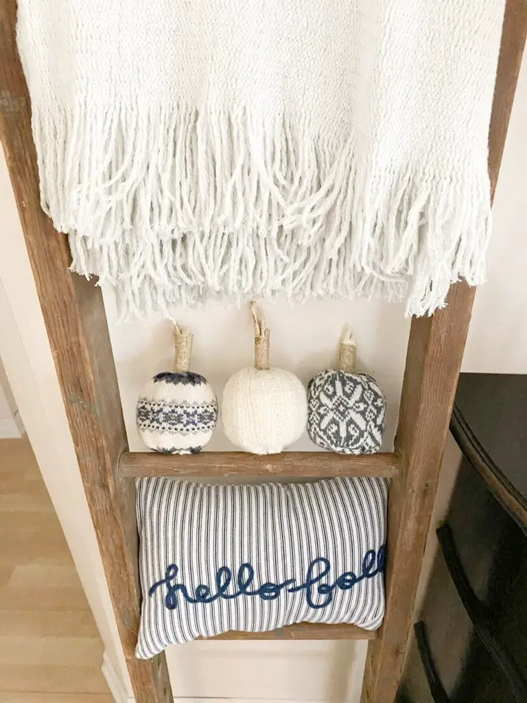 Photo shows the 3 completed DIY sweater pumpkins sitting on an antique ladder underneath a grey throw blanket and above a blue and white striped pillow that says "hello fall"