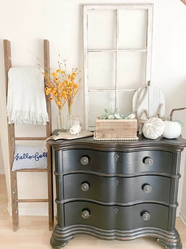 Entryway table vignette with muted fall decor. There is an old ladder with a blanket and pillow on it, an old window on the the table alongwith various fall decor items such as muted yellow flowers, white and white and gray patterned pumpkins, a wooden pumpkin sign with a wooden "hi" in front, and a wooden box with faux lambs ear and mini pumpkins inside.