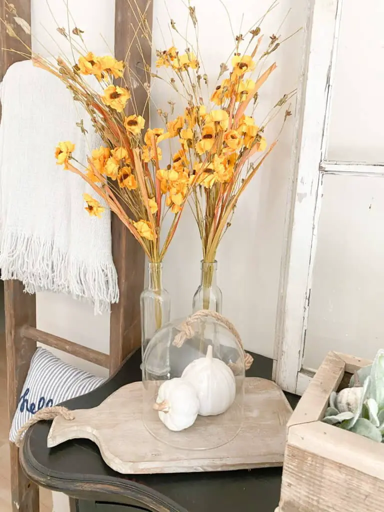 Some muted fall decor pieces, muted yellow flowers in glass bottles, a wood tray with 2 white pumpkins under a cloche.