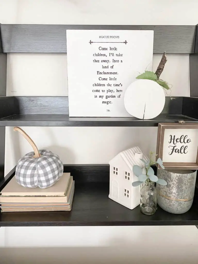 Black wall shelf with neutral and muted fall decor pieces including a gingham pumpkin, a wooden pumpkin, a fall sign, and a hocus pocus sign.