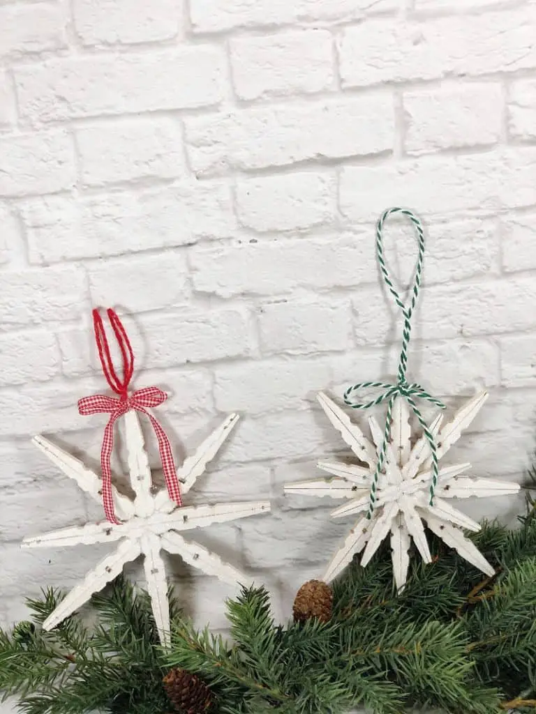 2 snowflake ornaments made out of clothespins and painted white. One has a red and white plaid ribbon and the other has a green and white striped ribbon. They are set against a white brick wall on top of a pine garland.