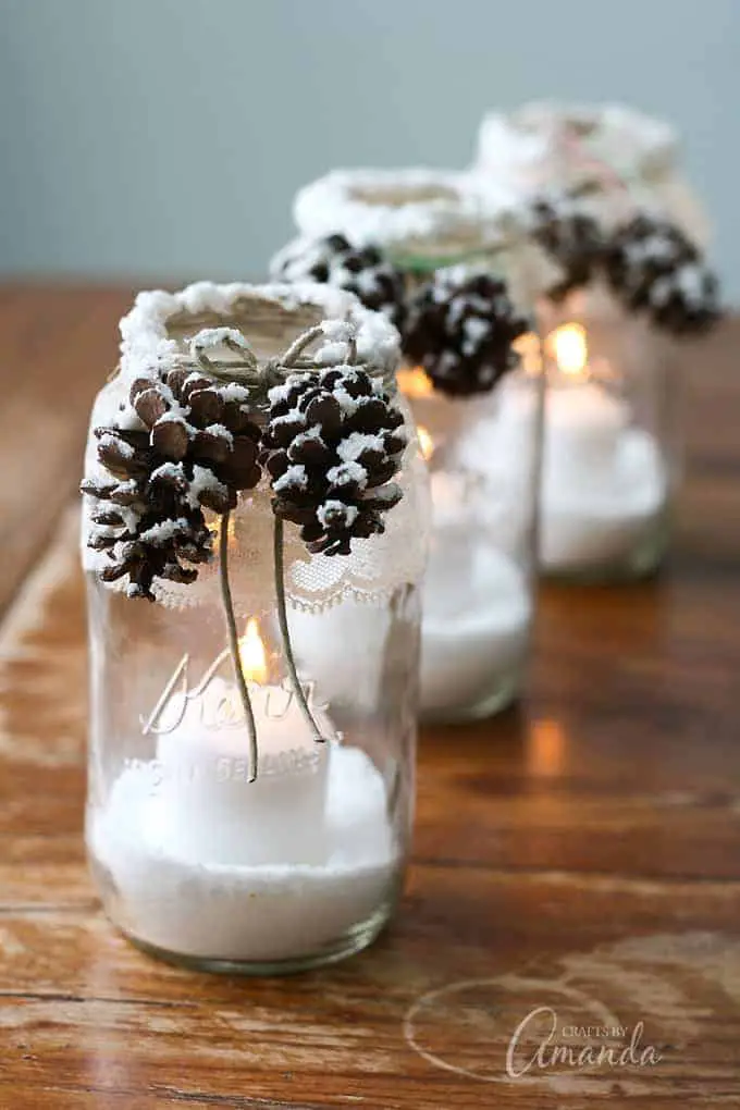 A set of 3 mason jars with fake snow and a candle inside and with lace wrapped around the top of the outside and some pinecones with fake snow attached to the front and a bow made from twine.