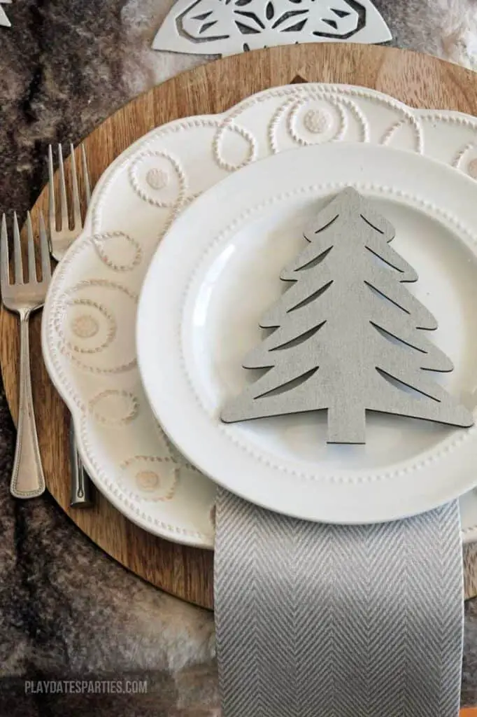 A neutral Christmas place setting. Includes a wood charger, 2 white plates with ornate detailing along the edges, 2 forks, a white and gray patterned napkin, and a gray wooden Christmas tree on the plates for decoration.