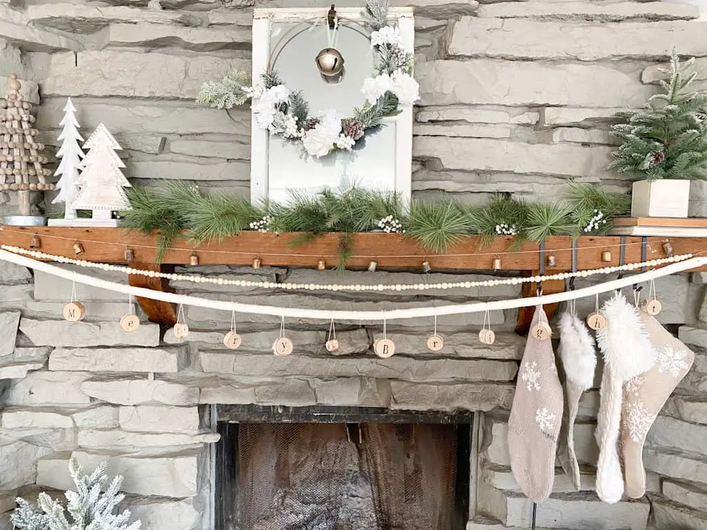 DIY Christmas Mantle Garlands. Image shws a fireplace decorated for Christmas with a garland, a mirror with a wreath, some faux pine trees, stockings, and 3 garlands. One if made from bells, one made from wood beads, and one made from pieces of wood with the words Merry & Bright on it.