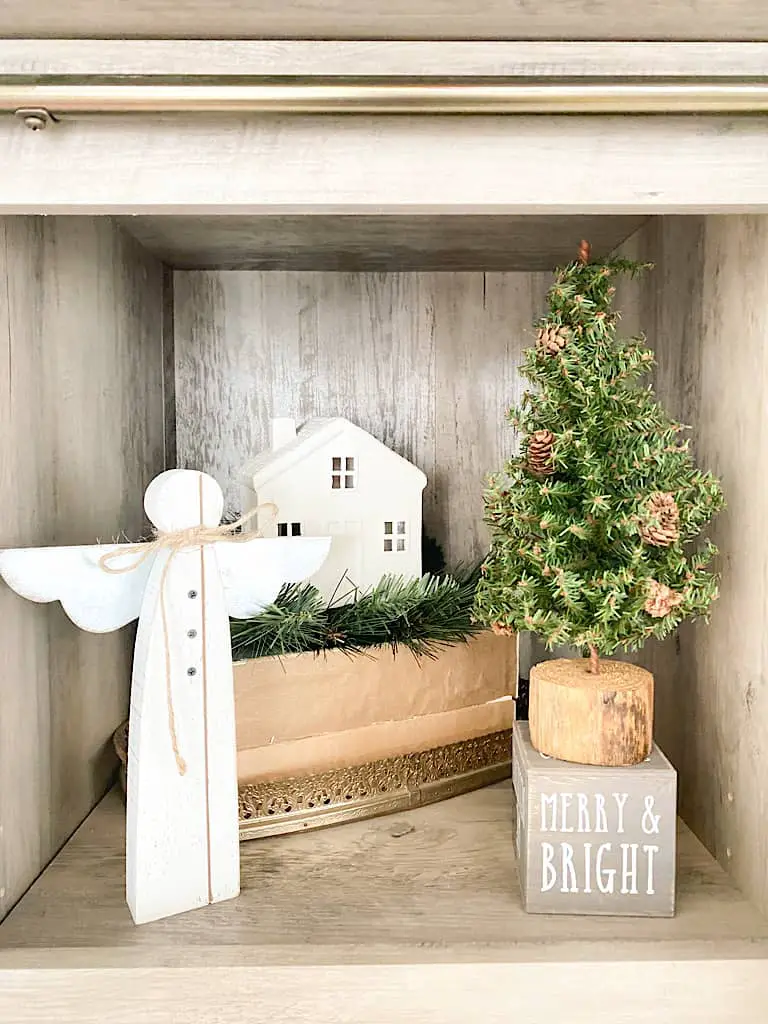 This shelf houses a little white wooden angel, a small mini pine tree with a wooden base on top of a gray wood block with the words "merry & bright" written in white on it. Behind that is a gold framed mirrored tray with a stack of 2 books on it. On top of those are some faux pine stems and a white ceramic house on top.