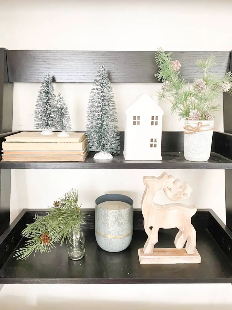 This black 2-tier shelf houses a Christmas vignette. There are some bottle brush trees on top of a stack of old books and one next to the stack of books. Next to that is a white ceramic house and then next to that is a small mason jar painted white with some twine tied around the top and there are some frosted pine branches sticking out from it. The bottom shelf has a small glass bottle that has a faux spruce stem hanging out of the top, a candle in a metal canister, and a wooden deer.