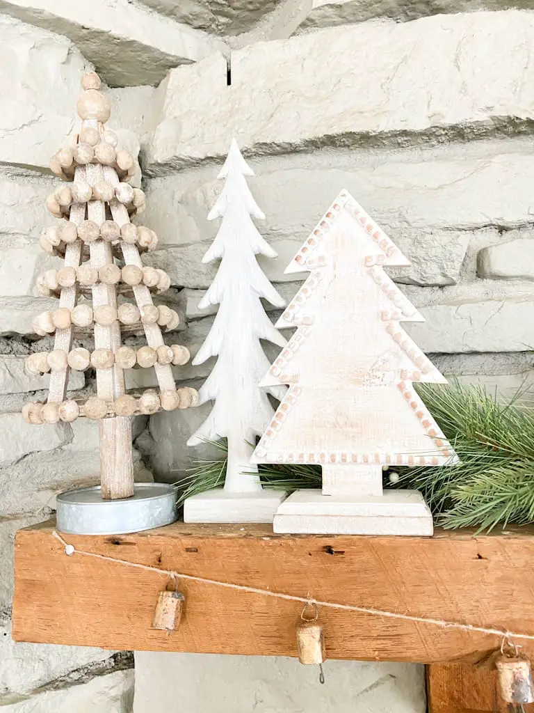 A set of 3 wood trees on a wooden mantle with some spruce branches behind them. One tree is a wood bead tree that is slightly whitewashed with a metal base. The other 2 are painted white and one is taller and skinner, the other is wider and has some wood beads outlining it.