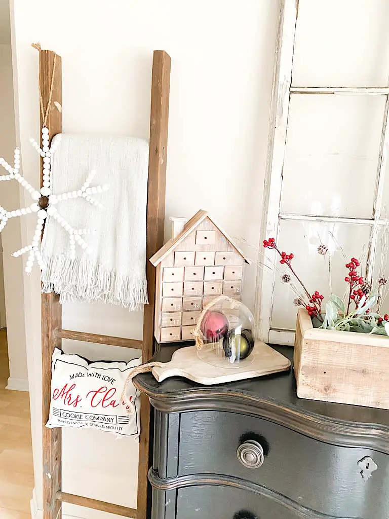 An old ladder with a gray throw blanket, a Mrs. Claus pillow, and a white wood bead snowflake hanging on it. Next to that is the edge of a black dresser with a wooden house advent calendar, a large red and a large green jingle bell under a glass cloche and on top of a wood paddle cutting board.