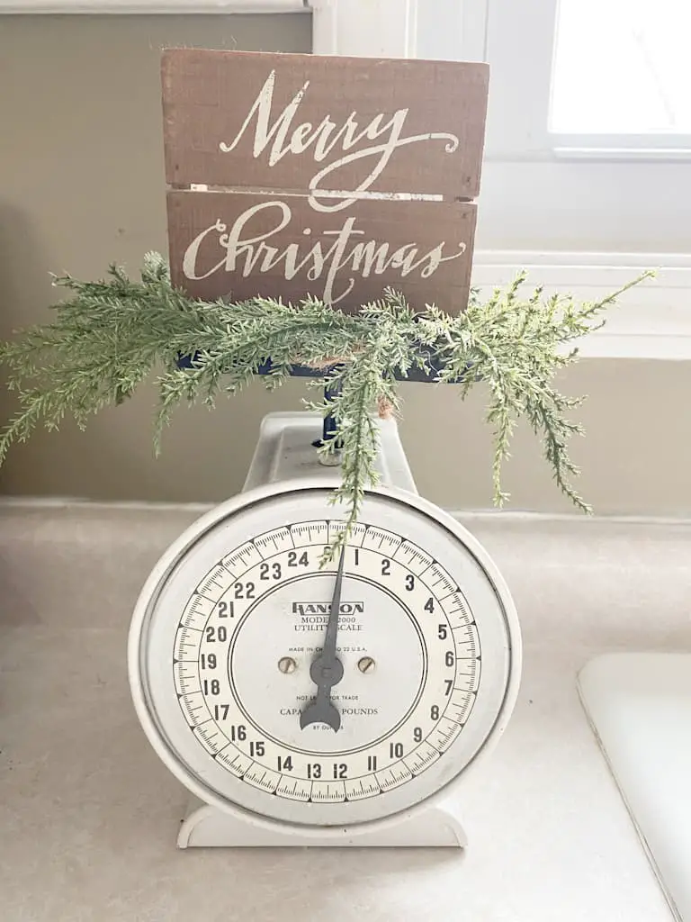 This white antique kitchen scale has a faux pine wreath sitting on top of it and a small wooden sign that says "Merry Christmas."