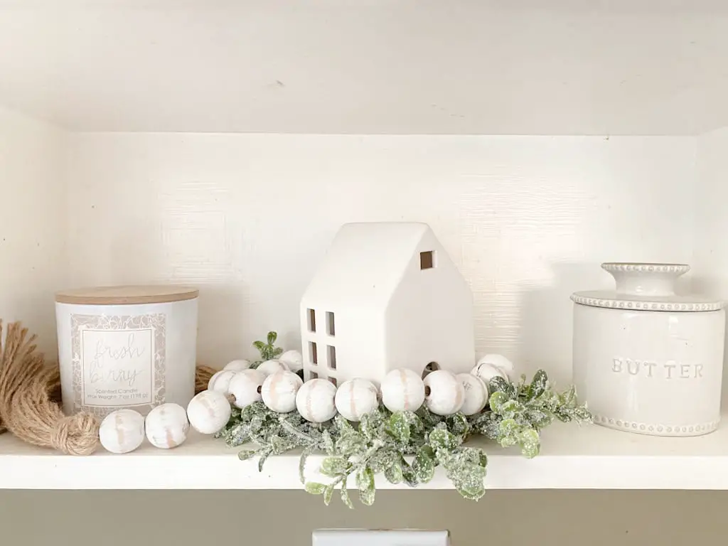 A shelf with a white jar candle with a wooden lid, a white ceramic house with a white wood bead garland wrapped around it and sitting on a small wreath of faux snowy leaves, and a small ceramic butter jar with a lid.