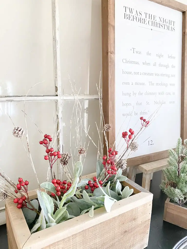 This wood box filled with faux lambs ear, faux red berry branches, and faux birch branches add lots of texture and a little pop of color to this neutral Christmas décor. Behind the box is an old window, a wood framed sign that has a passage from the Night Before Christmas sitting on a small wood stool and part of a faux pine tree is showing as well.