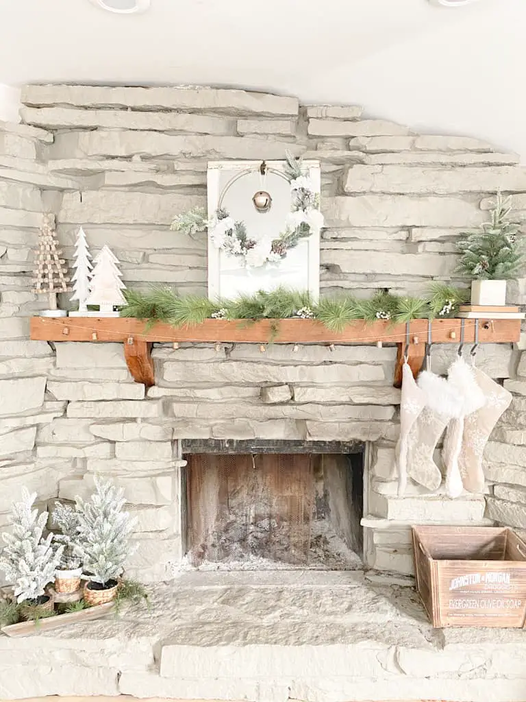 A fireplace with neutral Christmas decorations. There is an old window turned into a mirror with a hoop wreath and an old gold bell hanging from it, a set of 3 wood trees, a spruce garland, a faux pine tree in a box on top of 2 old books, 4 knit stocking hanging from black metal hooks, an antique bell garland, a wood box, and 3 mini flocked pine trees in different wicker baskets sitting on a wood tray filled with faux spruce branches.