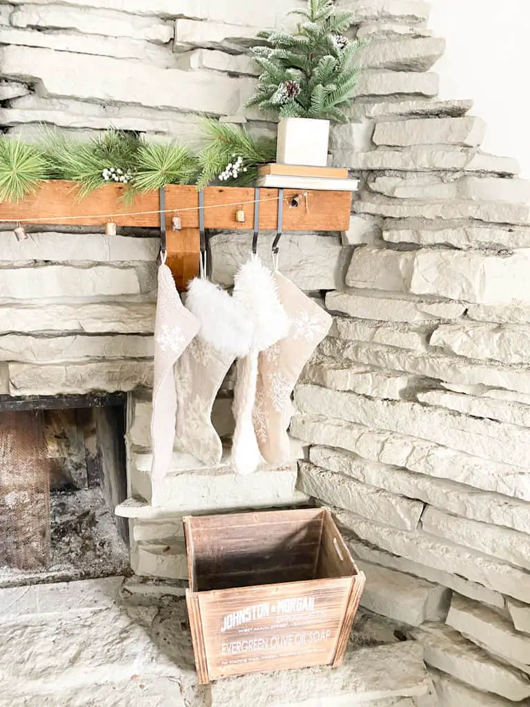 A set of 4 different knit stocking hanging from black metal hooks on a wood fireplace mantle. There is also a garland made from antique bells, a spruce garland, and a mini faux pine tree in a box on top of 2 old books. A wooden box sits below on the ledge of the stone fireplace.