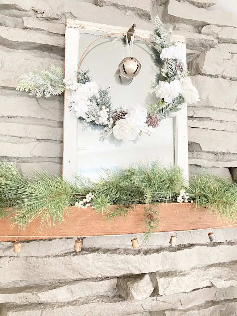 an old window turned into a mirror with a hoop wreath and an old gold bell hanging from it, a spruce garland with white berries mixed in, and an antique bell garland.