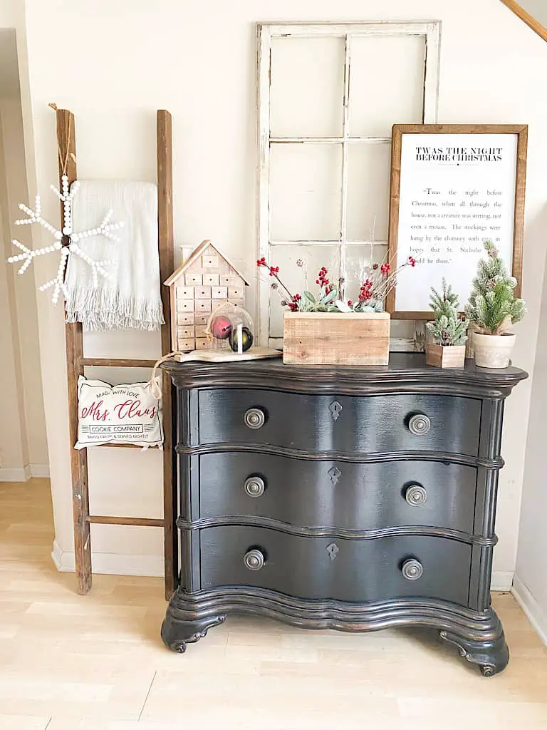 This black antique looking dresser houses a large antique window, a wood framed sign with a passage from The Night Before Christmas, 2 faux mini pine trees, wood box filled with faux lambs ear, faux red berry branches, and faux birch branches, 2 large vintage bells in red and green under a glass cloche sitting on a wood paddle cutting board, and a wooden house advent calendar. Next to the dresser is an old ladder with a gray throw blanket, a Mrs. Claus pillow, and a white wood bead snowflake hanging on it. This all adds warmth, texture, and a little color to these neutral Christmas decorations.