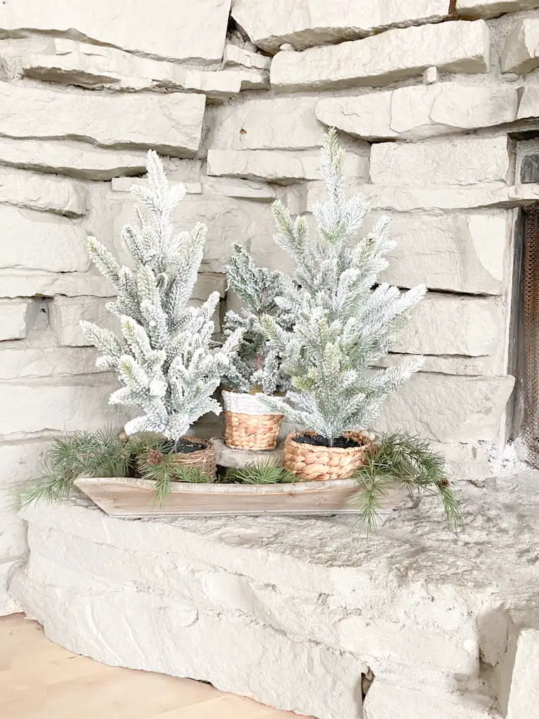 A set of 3 different flocked faux pine trees in assorted wicker baskets. They are sitting on a wood tray with faux spruce branches.